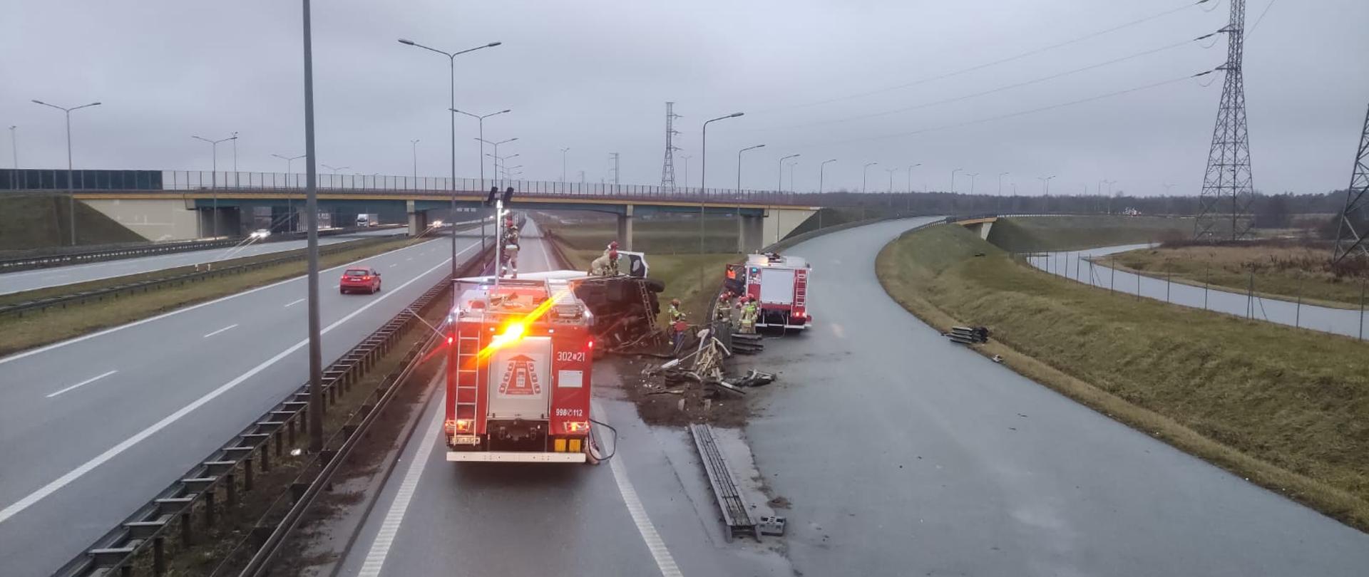 Zdjęcie przedstawia panoramę drogi S74 i wjazd na drogę ekspresową S7. Na pasie oddzielającym leży przewrócona ciężarówka a po obu stronach stoją samochody pożarnicze.