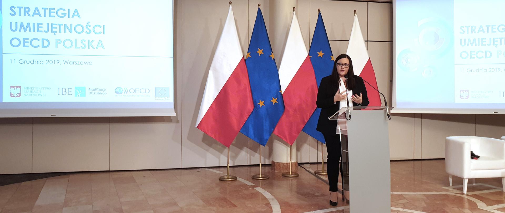 minister Małgorzata Jarosińska-Jedynak mówi do mikrofonu stojąc przed mównicą, za nią stoją flagi Polski i Unii Europejskiej, obok flag są ekrany z napisem "Strategia umiejętności OECD: Polska, 11 grudnia 2019 , Warszawa