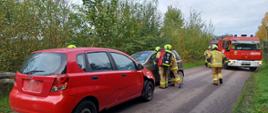 Zdjęcie przedstawia dwa samochody osobowe po zderzeniu stojące na drodze. Strażacy wykonują rozpoznanie. W tle samochód pożarniczy i drzewa.