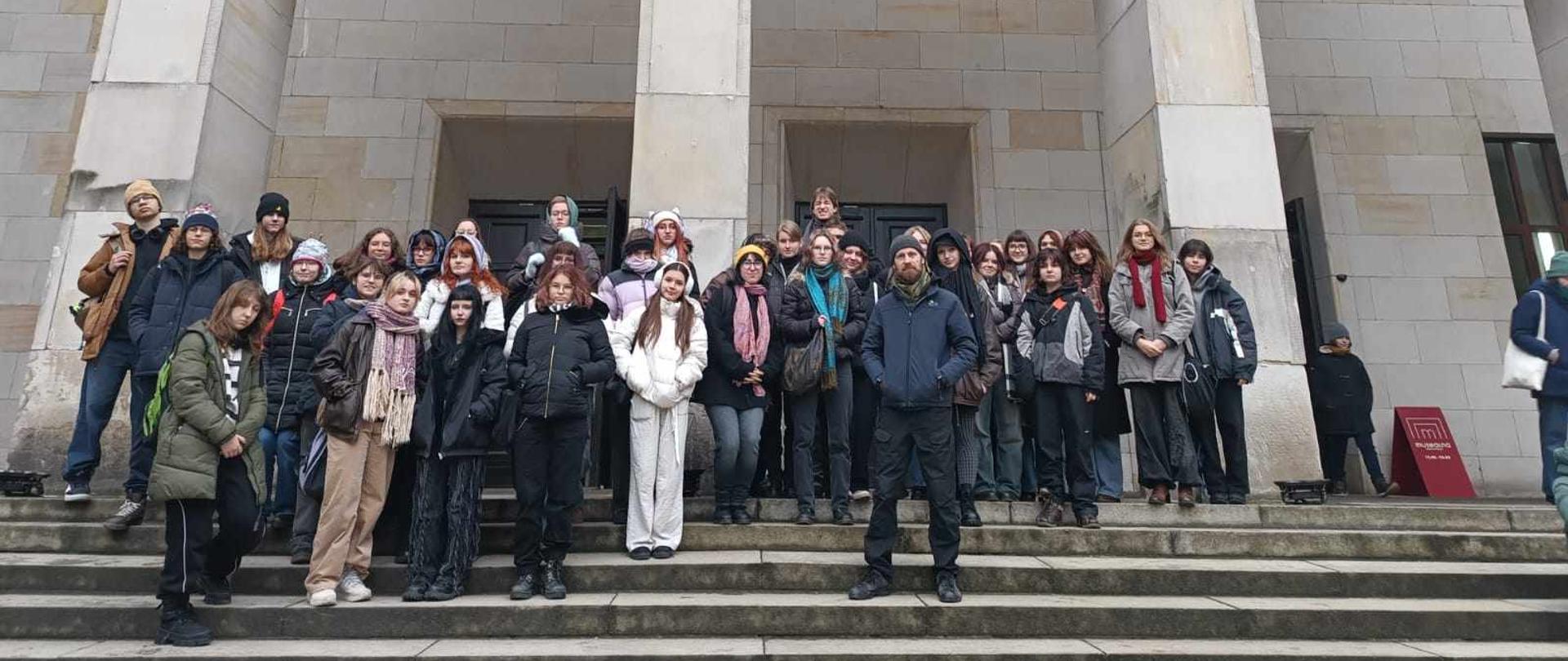 Zdjęcie grupowe, pozowane. Uczniowie i nauczyciel Wojciech Koryciński stoją przed wejściem do Muzeum Narodowego w Warszawie. Na jego frontowej ścianie znajduje się wielki, trzyczęściowy baner reklamujący wystawę obrazów J. Chełmońskiego od 27.09 do 26.01. Na banerze oprócz dat, napisu Józef Chełmoński reprodukcja pejzażu artysty ukazująca ptaka w locie nisko akwenu i roślinności.