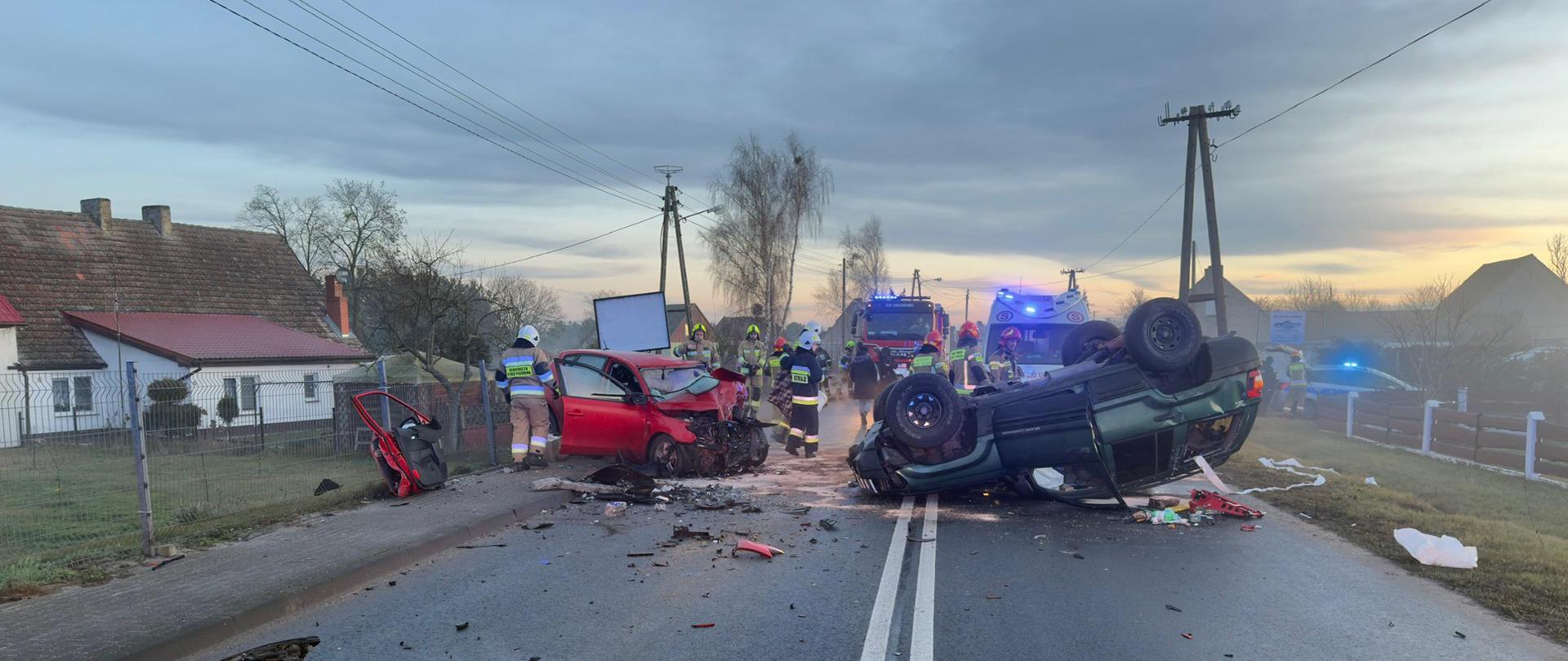 Fotografia przedstawia ujęcie wzdłuż jezdni. Na prawym pasie, w poprzek jezdni leży na dachu samochód osobowy w kolorze zielonym, dookoła pojazdu leżą elementy karoserii, widoczny płat poduszki powietrznej kierowcy. Częściowo na lewym pasie, częściowo na poboczu stoi czerwony samochód osobowy z rozbitym przodem, przy pojazdach pracują strażacy.