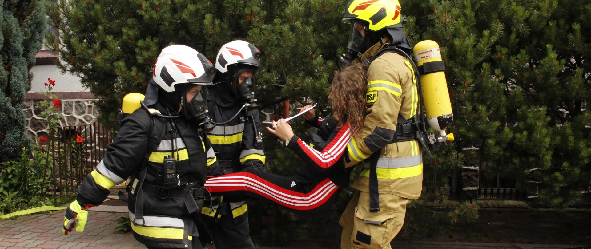 Zdjęcie przedstawia 3 druhów Ochotniczej Straży Pożarnej wynoszących poza budynkiem osobę poszkodowaną