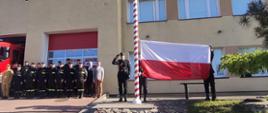 Na zdjęciu widać strażaków, pojazd strażacki (czerwony). Ze strażakami stoi Starosta Kętrzyński oraz kierownik Powiatowego Centrum zarządzania Kryzysowego. Poczet Flagowy przygotowany do wciągnięcia Flagi na maszt. Flaga rozłożona trzymana przez strażaka.