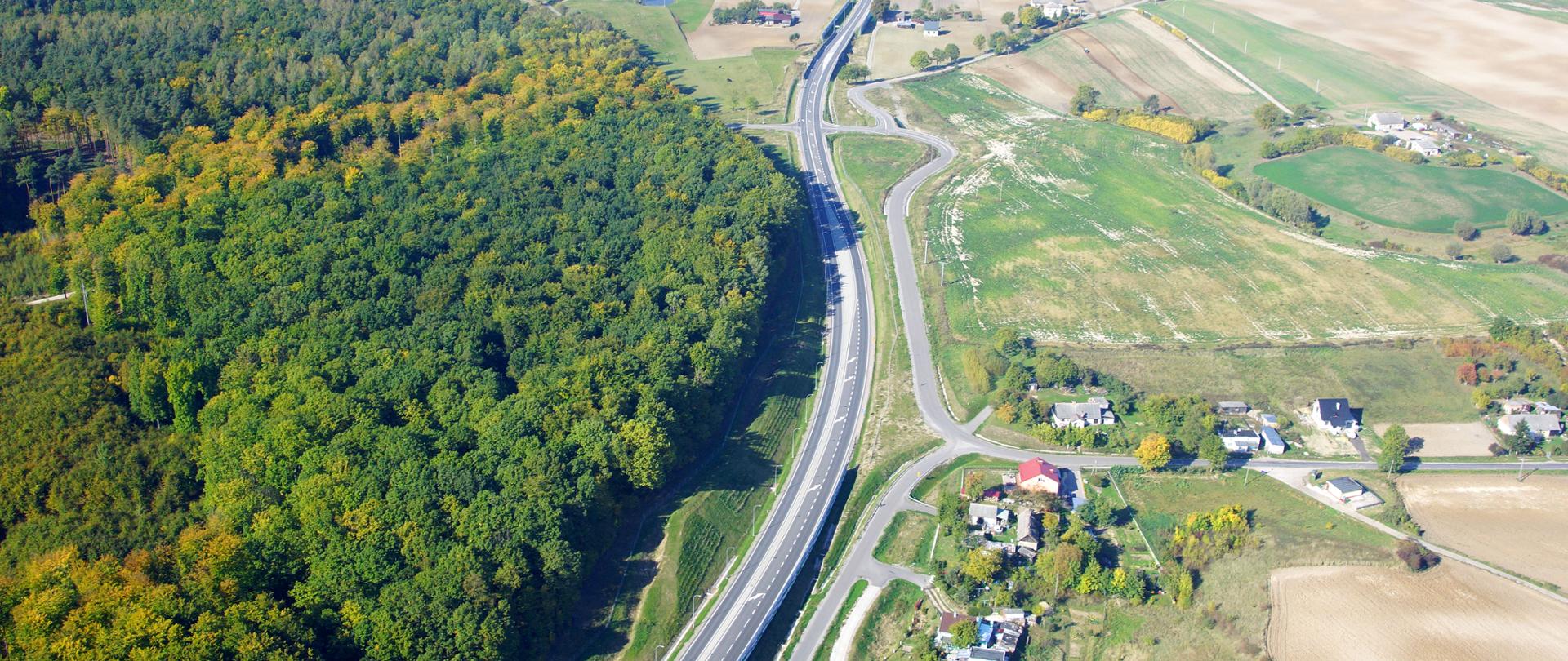 Zdjęcie przedstawia widok z lotu ptaka na drogę krajową obok drogi po lewej stronie las po prawej krajobraz rolniczy z zabudowaniami miejscowości.