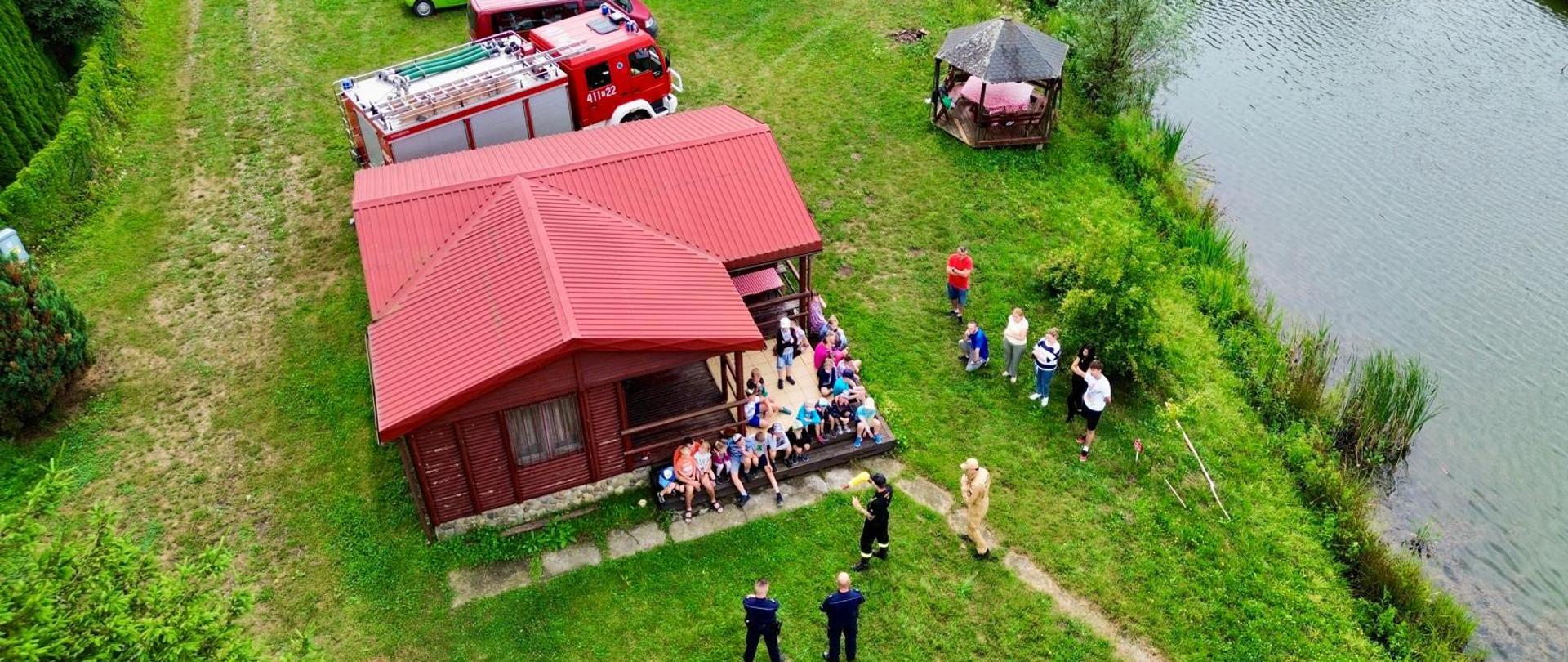 Kolorowa fotografia wykonana z lotu ptaka (dron) na zewnątrz w pochmurny dzień nad akwenem. Przedstawia strażaków w ubraniu służbowym, policjantów oraz dzieci które słuchają prelekcji na temat bezpiecznego wypoczynku nad wodą. Obok dzieci znajdują się opiekunowie. 