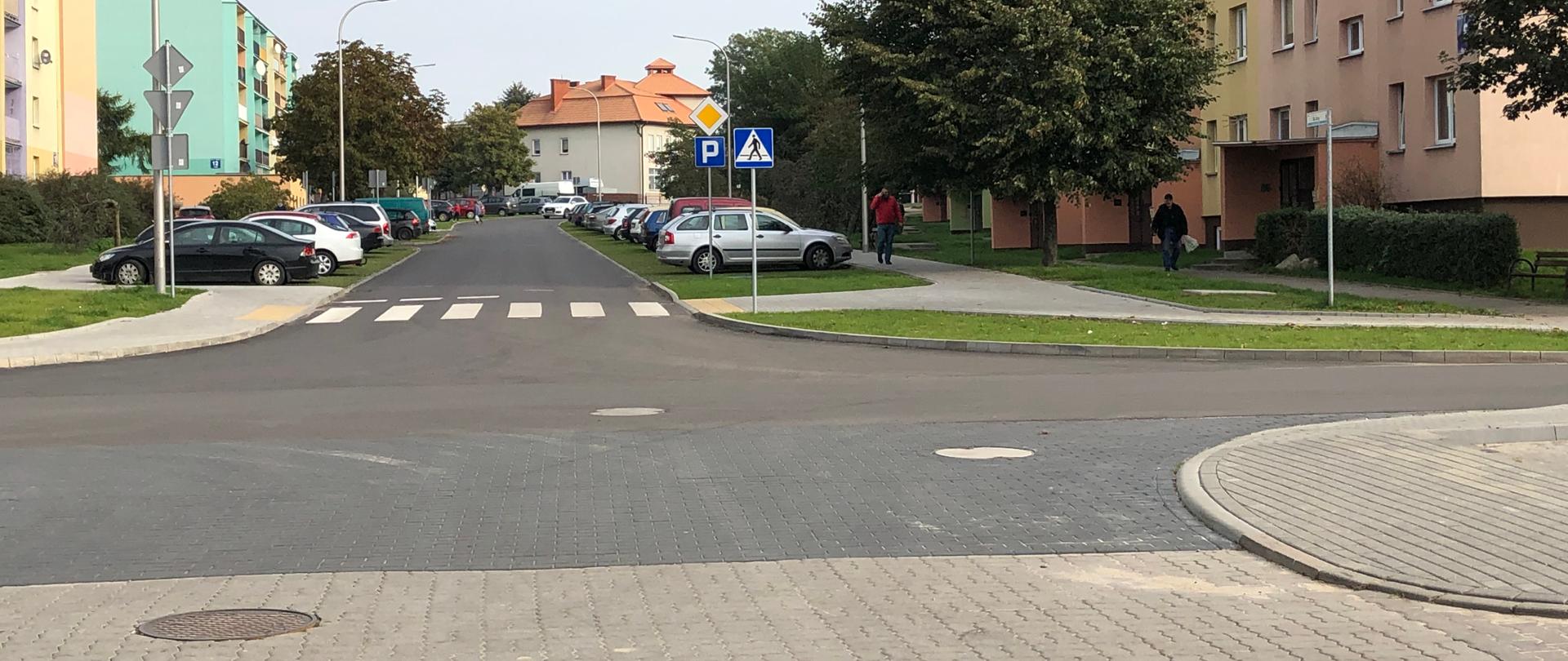 Na zdjęciu znajduje się droga, która została wyremontowana. W tle widać samochody oraz budynki.