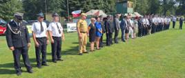 Na płycie boiska trwa ceremonia otwarcia zawodów. Na przeciwko ustawionych drużyn, w szeregu stoja zaproszeni goście. Wśród nich są strażacy, przedstawiciele władz samorządowych oraz komicja sędziowska. Przez mikrofon przemawia burmistrz Gminy Rawicz. W tle drzewa, namioty i budka sędziowska. 