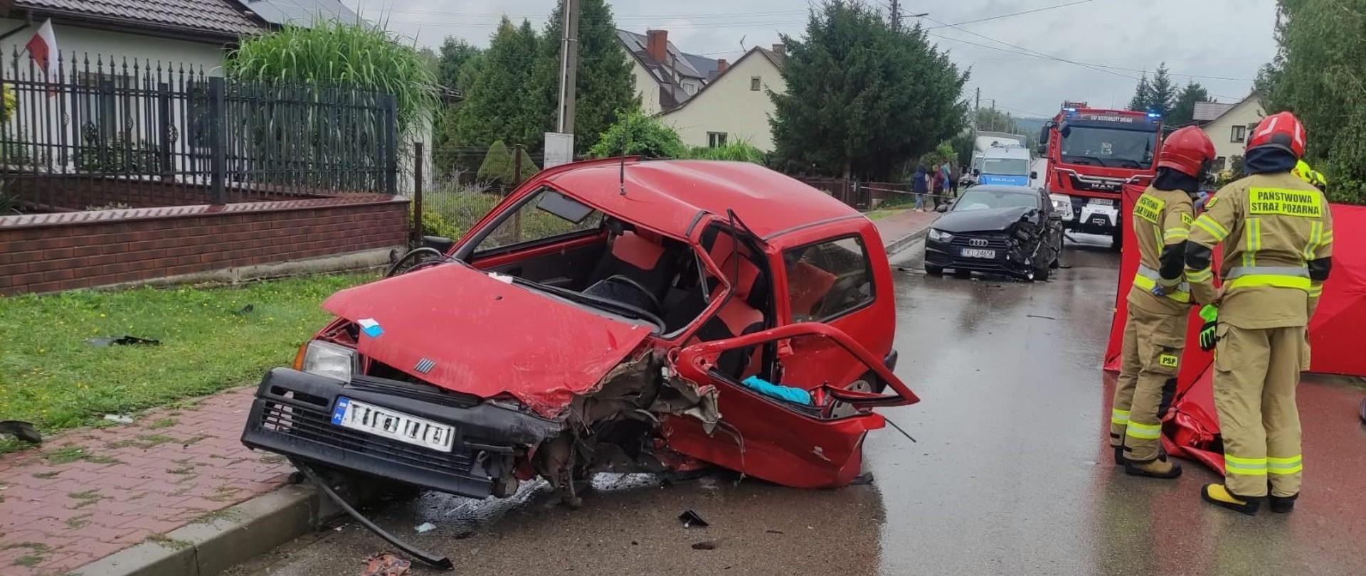 Zdjęcie przedstawia drogę powiatową. Na pierwszym planie jeden z pojazdów biorących udział w wypadku. Po prawej stronie dwóch ratowników stojących na tle czerwonego parawanu osłaniającego. W głębi stoi drugi samochód z wypadku a za nim wóz gaśniczy. 