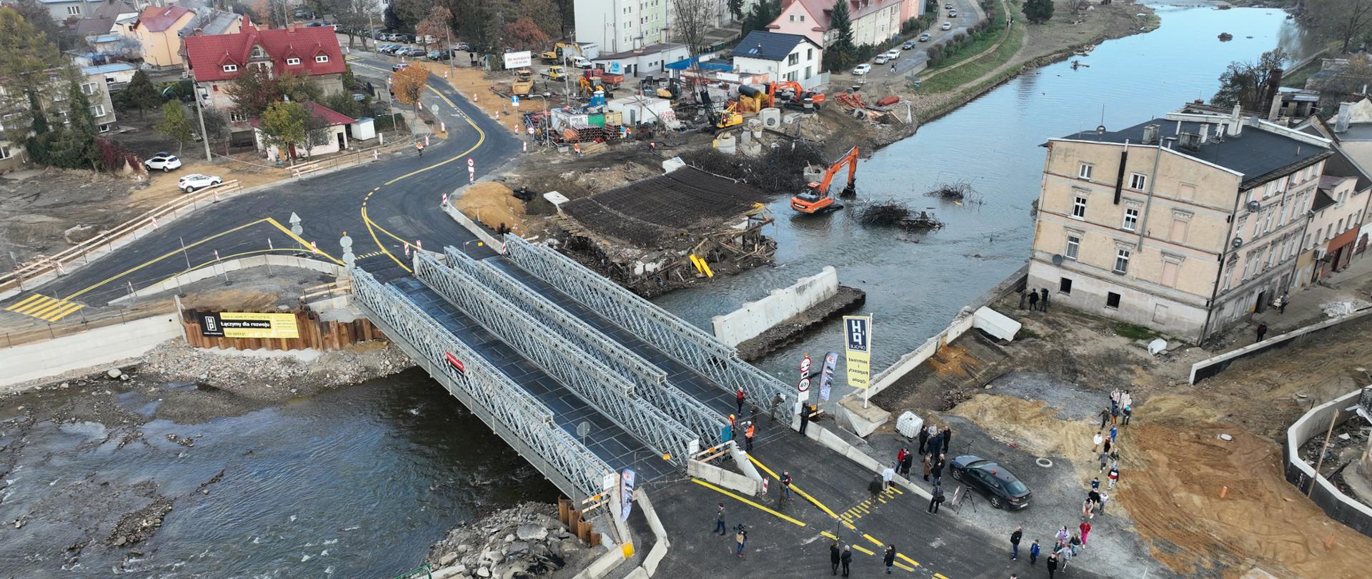 Zdjęcie przedstawia tymczasowy most na drodze krajowej nr 40 w Głuchołazach. Most jest wykonany z metalowych elementów i rozciąga się nad rzeką. Na moście i wokół niego znajdują się ludzie, prawdopodobnie uczestniczący w jego otwarciu lub przeprowadzający inspekcję. W tle widać koparkę i inne maszyny budowlane, a także znaki drogowe i oznaczenia na jezdni. Na dalszym planie widoczne są budynki mieszkalne oraz tereny zielone, co sugeruje, że most jest częścią większego projektu budowlanego lub remontowego w okolicy.