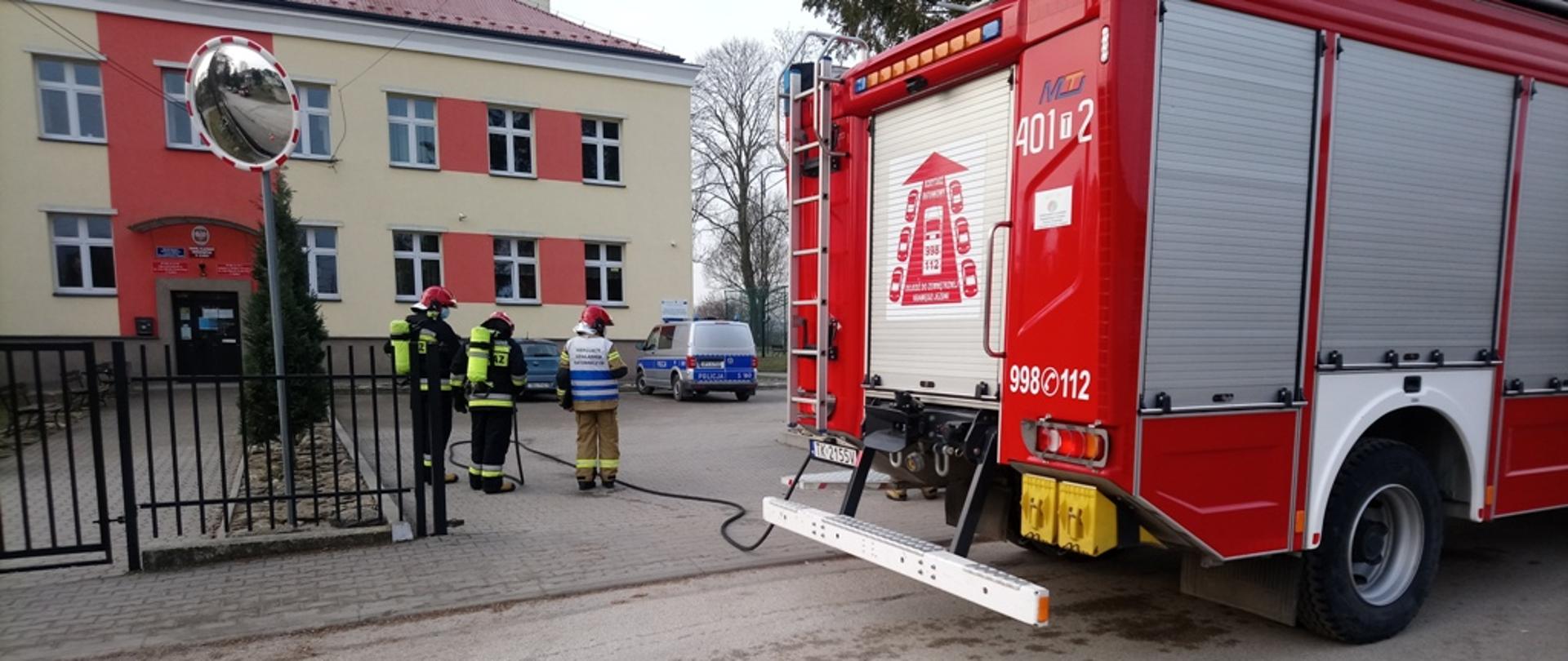 Na zdjęciu trzech strażaków zabezpieczających miejsce akcji, po prawej stronie zdjęcia pojazd pożarniczy.