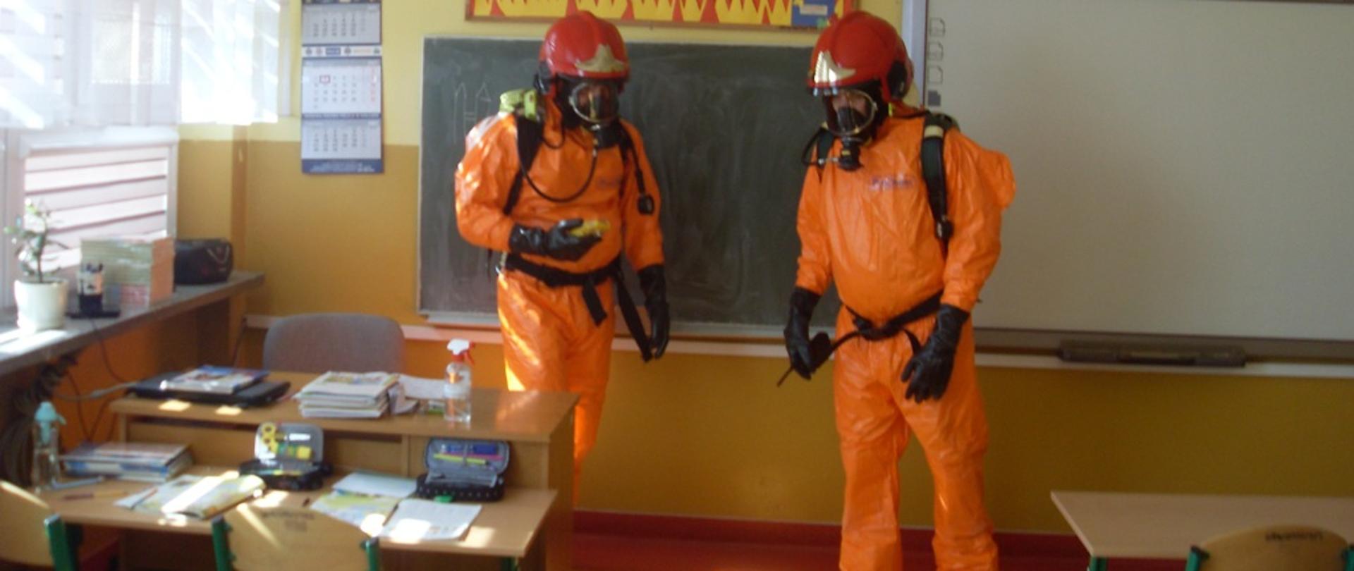 Zdjęcie przedstawia dwóch strażaków w ubraniu ochrony chemicznej, dokonujący pomiaru miernikiem wielogazowym jednego z pomieszczeń klasowych.