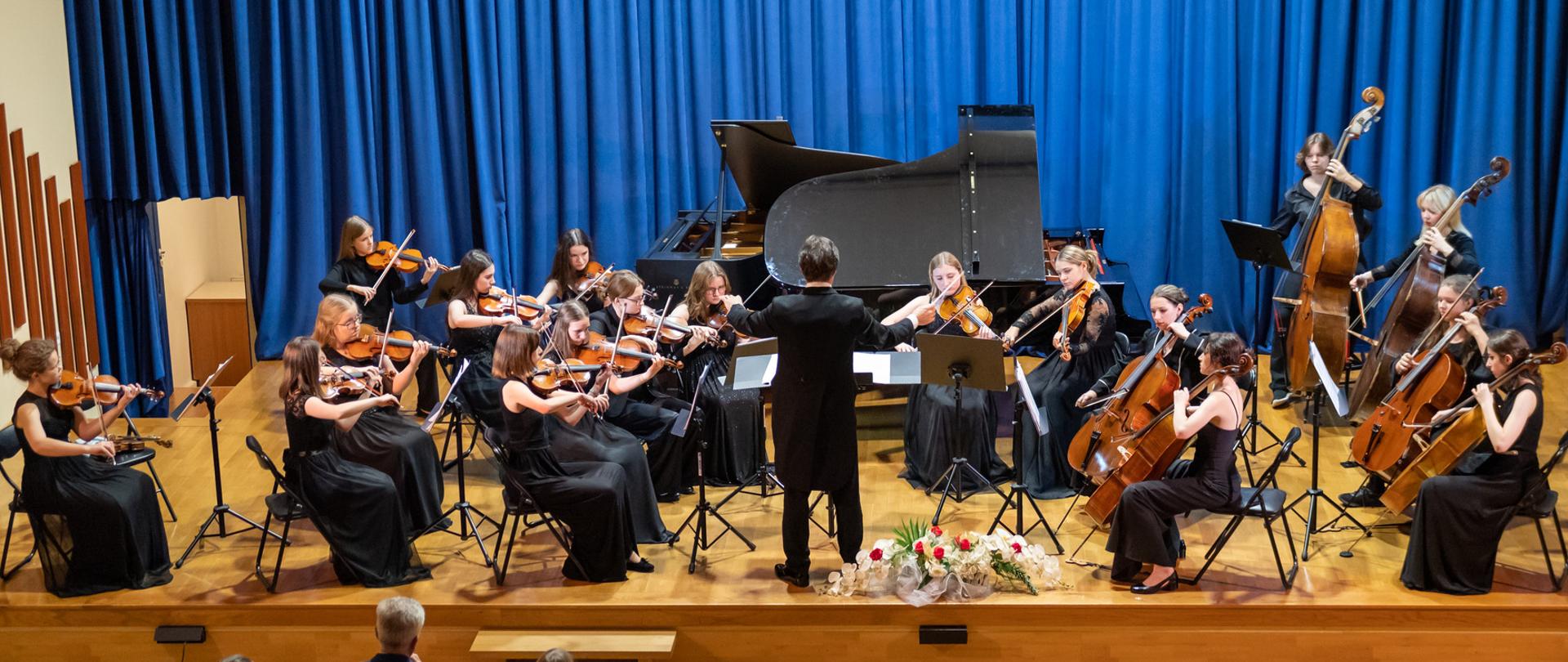 orkiestra kameralna grająca na instrumentach smyczkowych podczas koncertu na scenie, przed nią dyrygent, na scenie również bukiet kwiatów, przed sceną publiczność, tle niebieska kotara