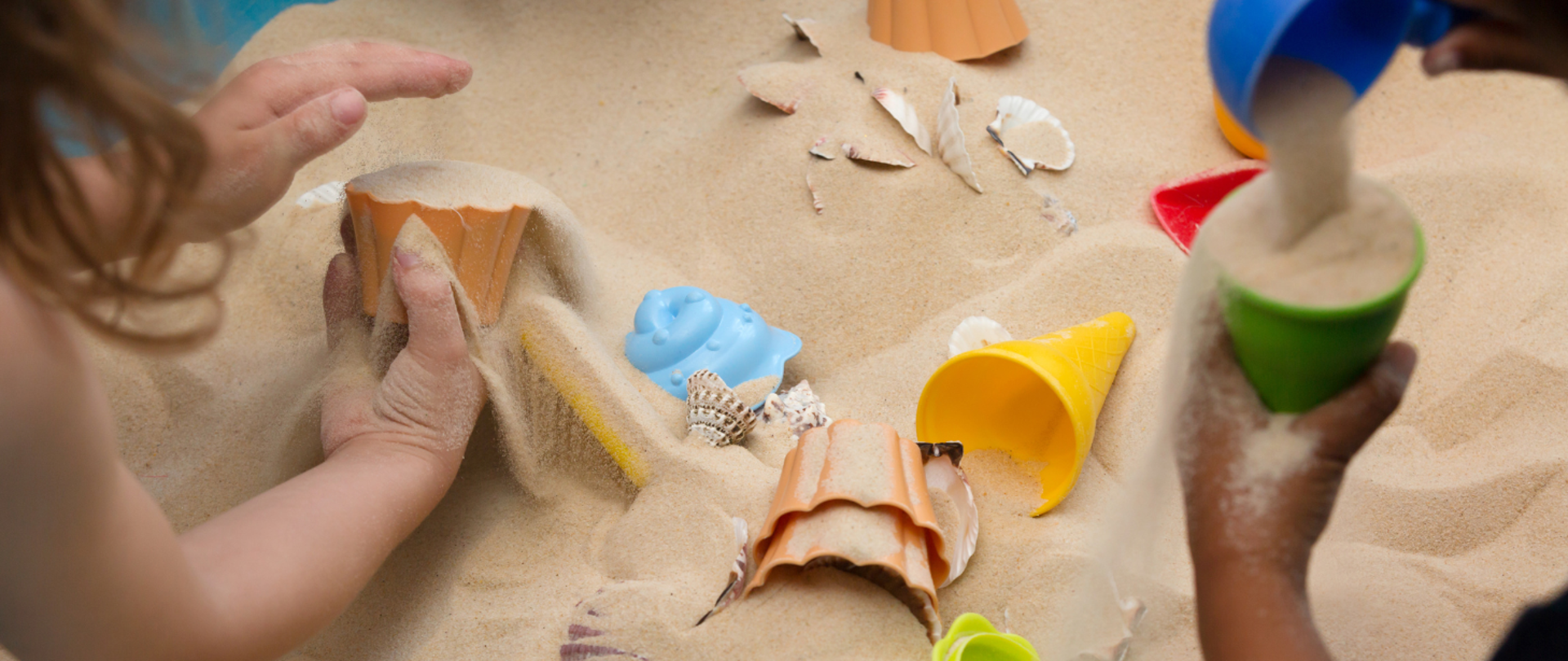 Zdjęcie przedstawia piasek na plaży i ręce dzieci bawiące się w piasku przy użyciu foremek. W piasku dodatkowo prócz foremek rozrzucone są muszelki.