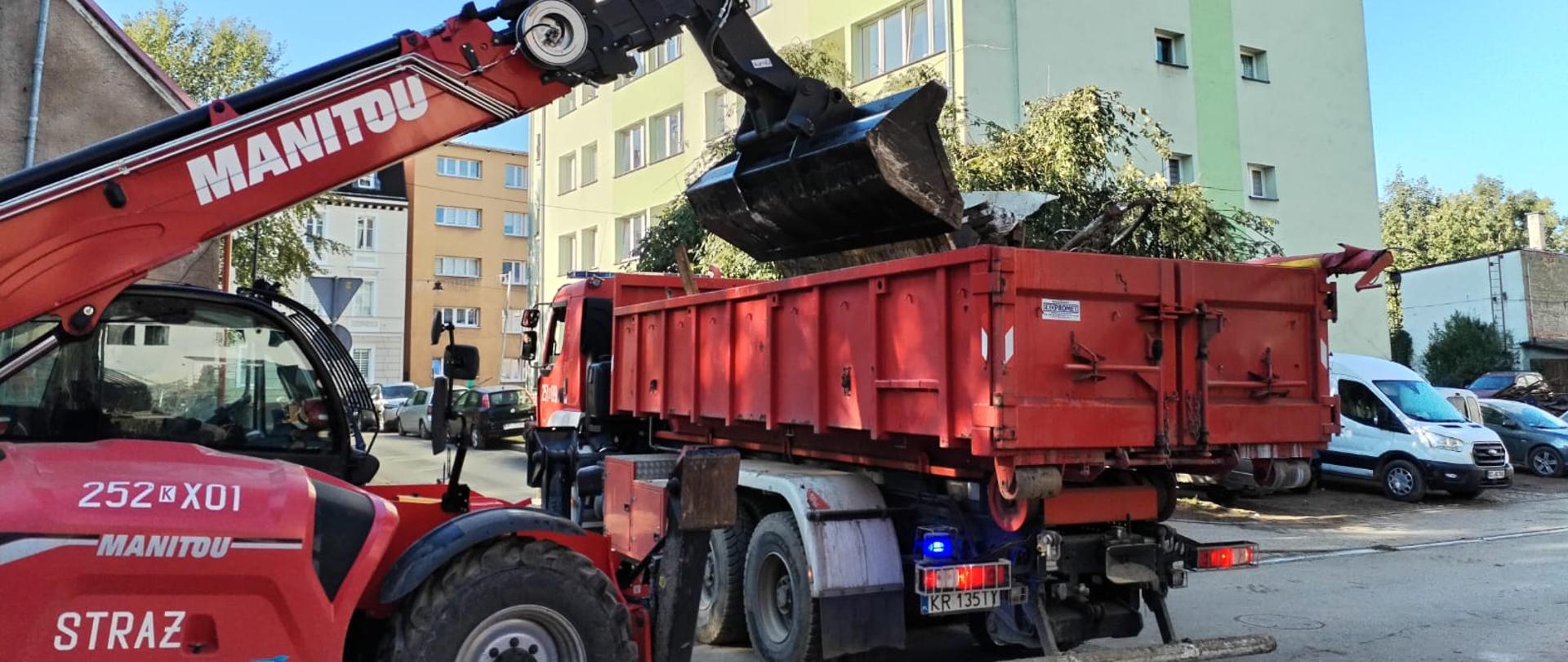 Samochód ciężarowy z kontenerem do którego gruzownik zrzuca gałęzie, w głębi widoczne bloki mieszkalne