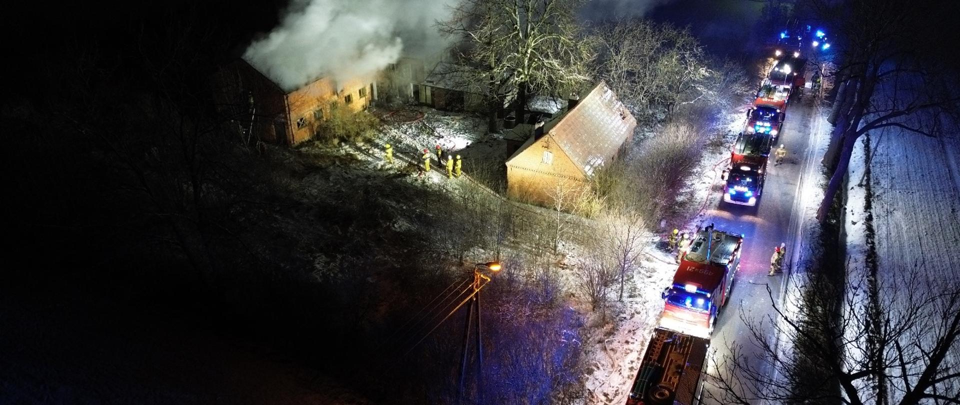 Zdjęcie wykonane z drona obrazujące pojazdy pożarnicze ustawione na drodze w kolumnie. Pojazdy mają włączone sygnały błyskowe. Trwa akcja gaśnicza. 