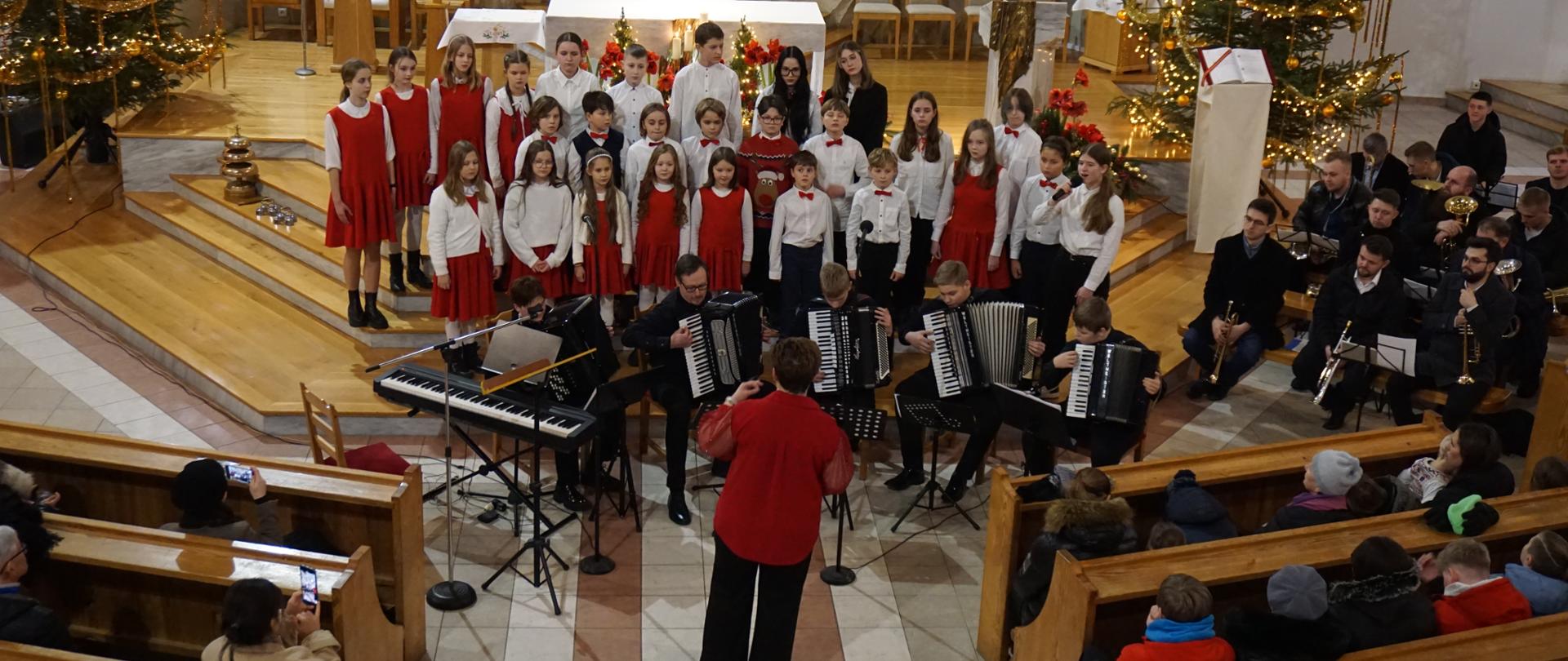 Przed ołtarzem w kościele NMP Królowej Polski w Wejherowie chór śpiewa kolędę. Przed na siedząco grają akordeoniści (kwintet). Dyrygent Ewa Rocławska dyryguje. Po bokach widać ławki i siedzącą publiczność.