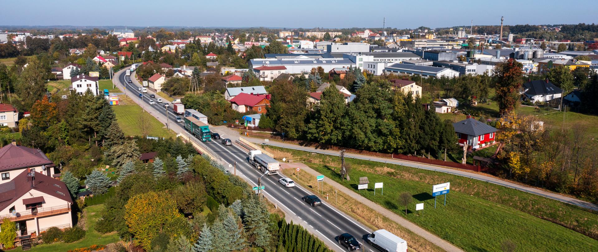 Widok z lotu ptaka na drogę krajową numer 9. Widać auta jadące drogą. Świeci słońce, po obu stronach drogi widać chodniki i rowy oraz zieleń dookoła drogi.