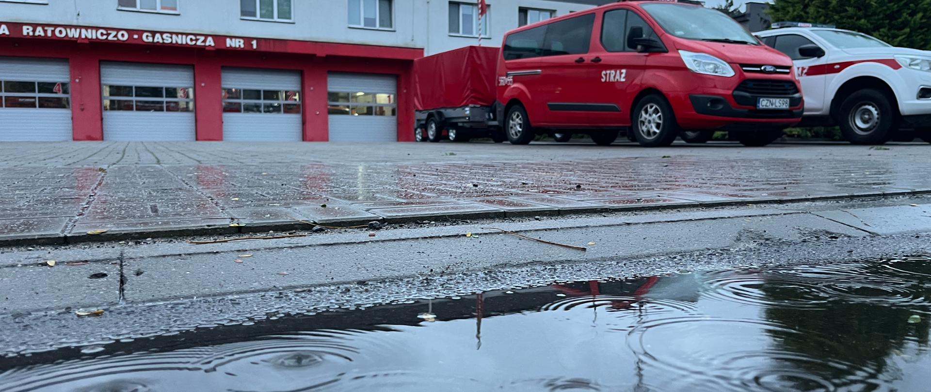 Na pierwszym polanie kałuża wody, pada deszcz. Dalej pojazdy pożarnicze, w dali fragment strażnicy - widok na wrota garażowe.