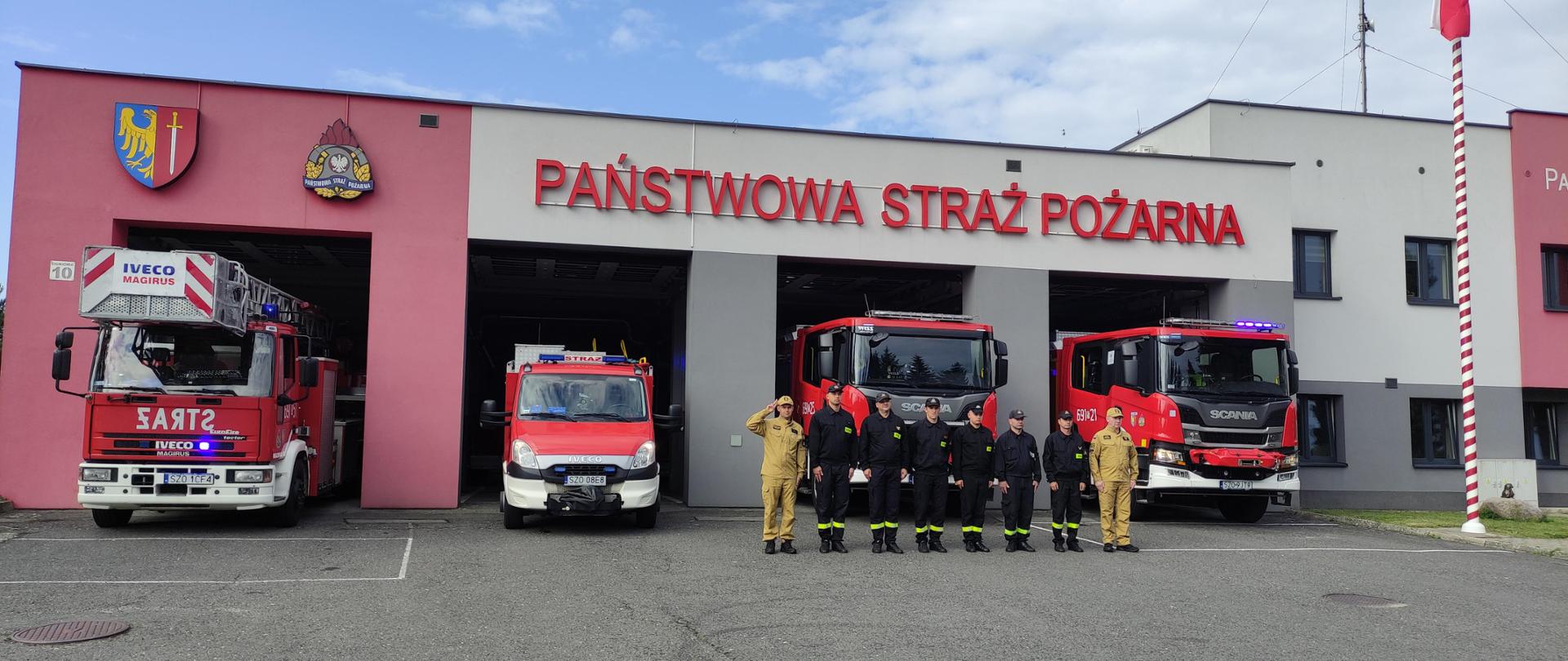 Grupa strażaków stoi w szeregu oddając hołd w rocznice 80 lecia Powstania Warszawskiego, w tle widać cztery wozy strażackie w kolorze czerwonem i budynek komendy państwowej straży pożarnej w Żorach.