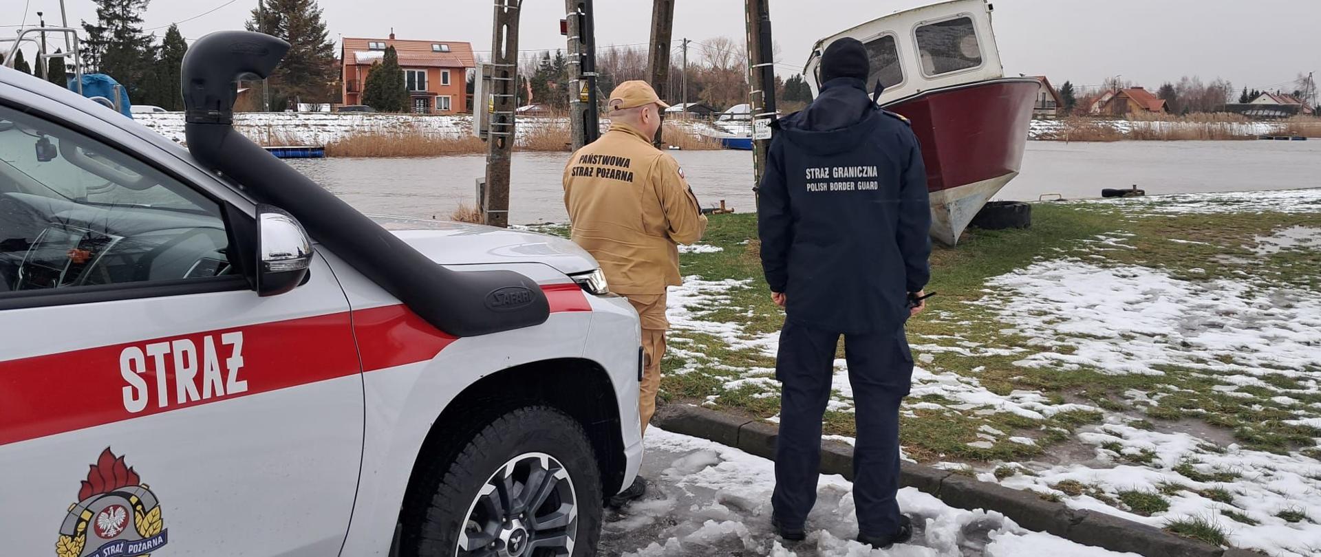 Z lewej strony widać samochód strażacki na środku stoi dwóch funkcjonariuszy. W oddali koryto rzeki, dużo wody.