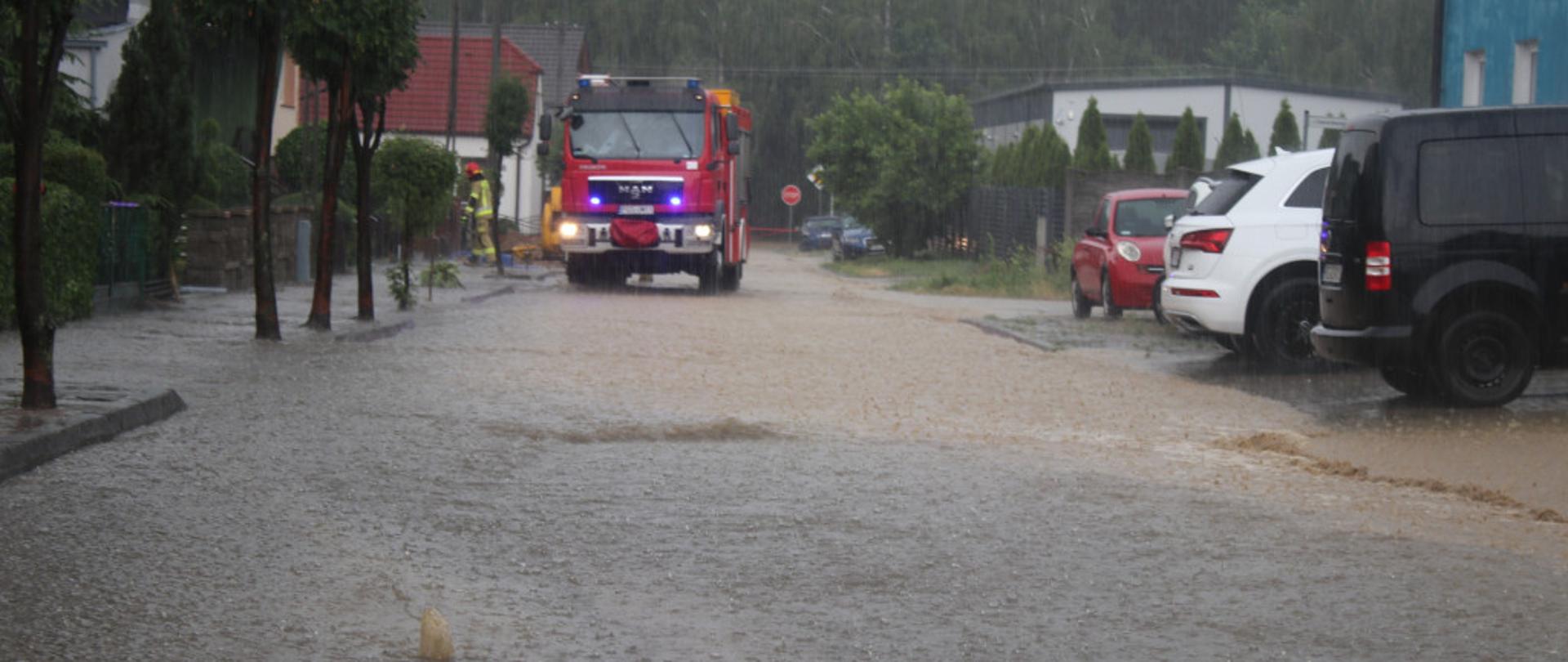 Na zdjęciu widać zalaną ulicę, stojące samochody. W tle znajduje się samochód pożarniczy podczas prowadzenia działań ratowniczych.