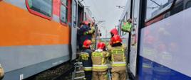 Zdjęcie przedstawia dwa pociągi pasażerskie na sąsiadujących torach. Między wagonami widać czterech strażaków w ubraniach specjalnych z czerwonymi hełmami na głowach, którzy przy użyciu podestu ratowniczego pomagają pasażerom przejść z jednego pociągu do drugiego.