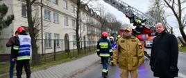 W tle strażacy prowadzą akcję ratowniczą z użyciem samochodu specjalnego - drabiny - usuwanie skutków silnego wiatru - zerwanie dachu. Na pierwszym planie stoi Zastępca Mazowieckiego Komendanta Wojewódzkiego PSP z Wojewodą Mazowieckim.