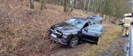 Fotografia przedstawia samochód osobowy marki mercedes w kolorze czarnym stojący w przydrożnym rowie, po prawej stronie kadru widoczni strażacy. W pojeździe są pootwierane drzwi kierowcy i tylna klapa bagażnika.