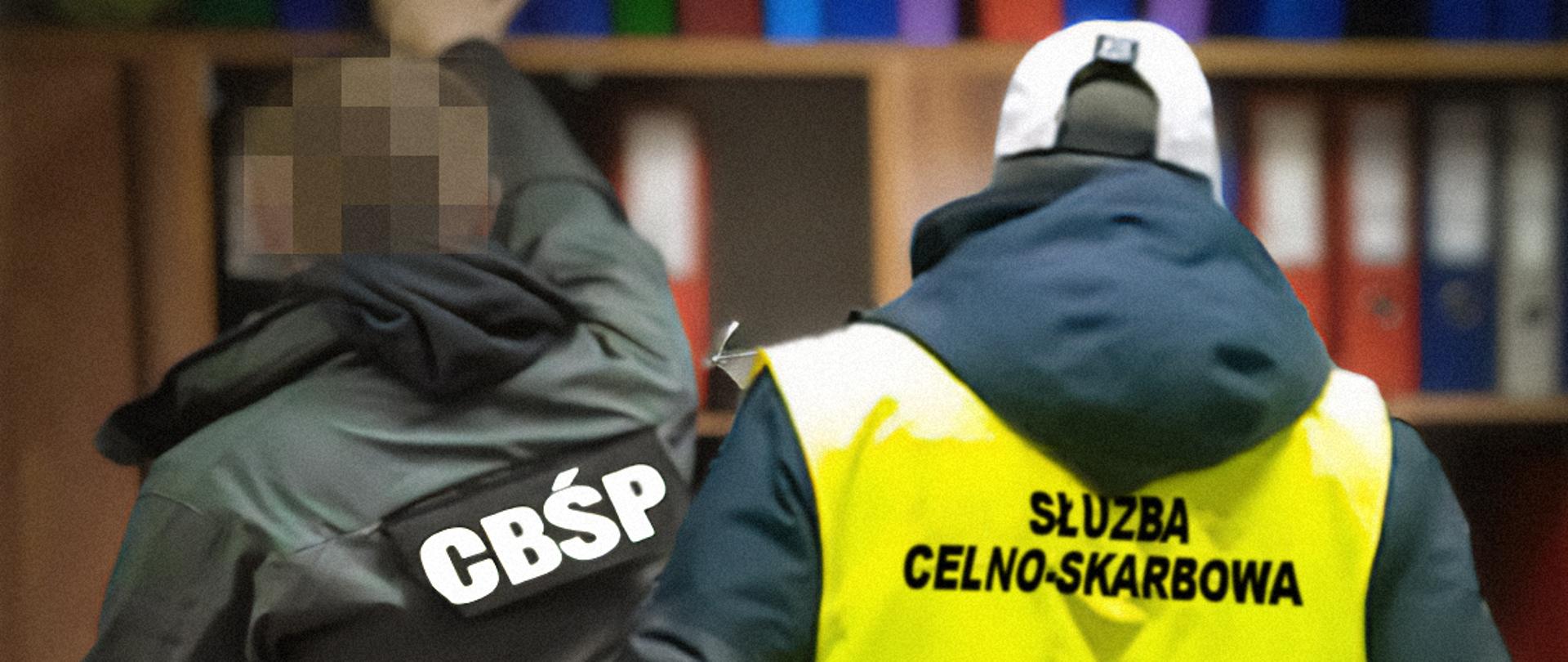 Na zdjęciu mężczyzna w kamizelce z napisem Służba Celno-Skarbowa oraz mężczyzna w kurtce z napisem CBŚP na tle regał z segregatorami.