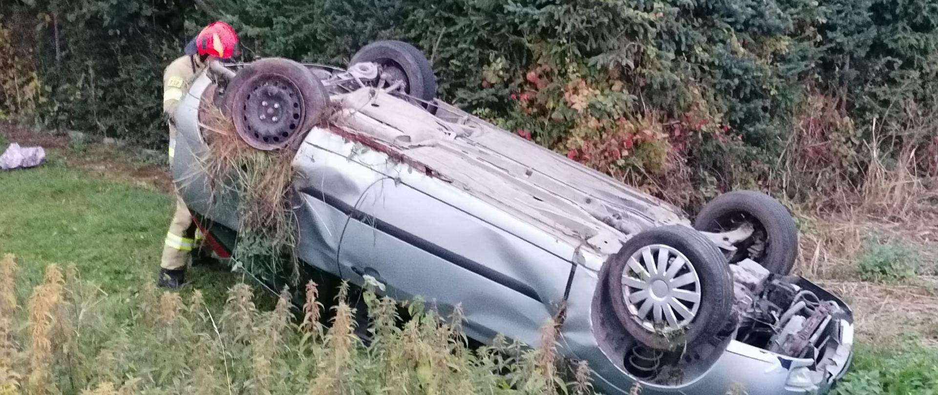 Srebrne auto osobowe leży na dachu na poboczu drogi, przy nim strażak w czerwonym hełmie.