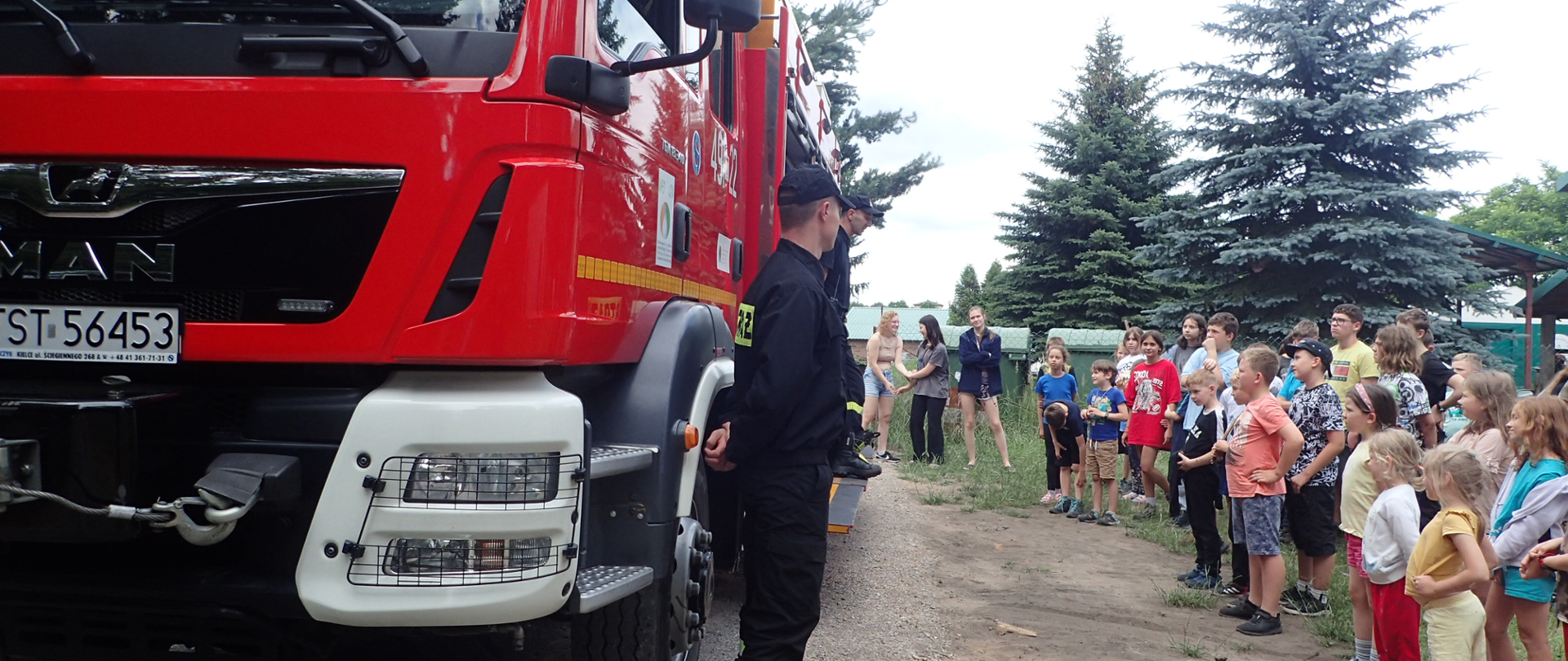 Po lewej stronie stoi wóz bojowy Państwowej Straży Pożarnej oraz strażacy skierowani w stronę grupki małych dzieci stojących po prawej stronie zdjęcia