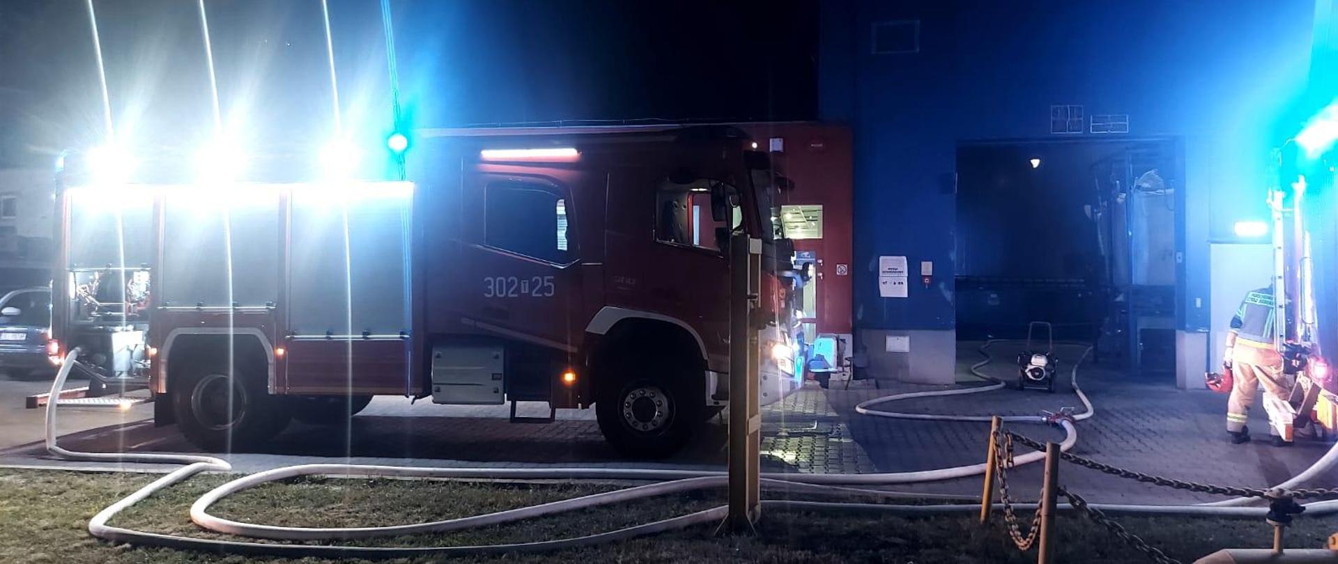 Zdjęcie przedstawia samochód pożarniczy z rozwiniętymi linami gaśniczymi stojący przed otwartą bramą wjazdową do budynku hali gdzie doszło do pożaru.
