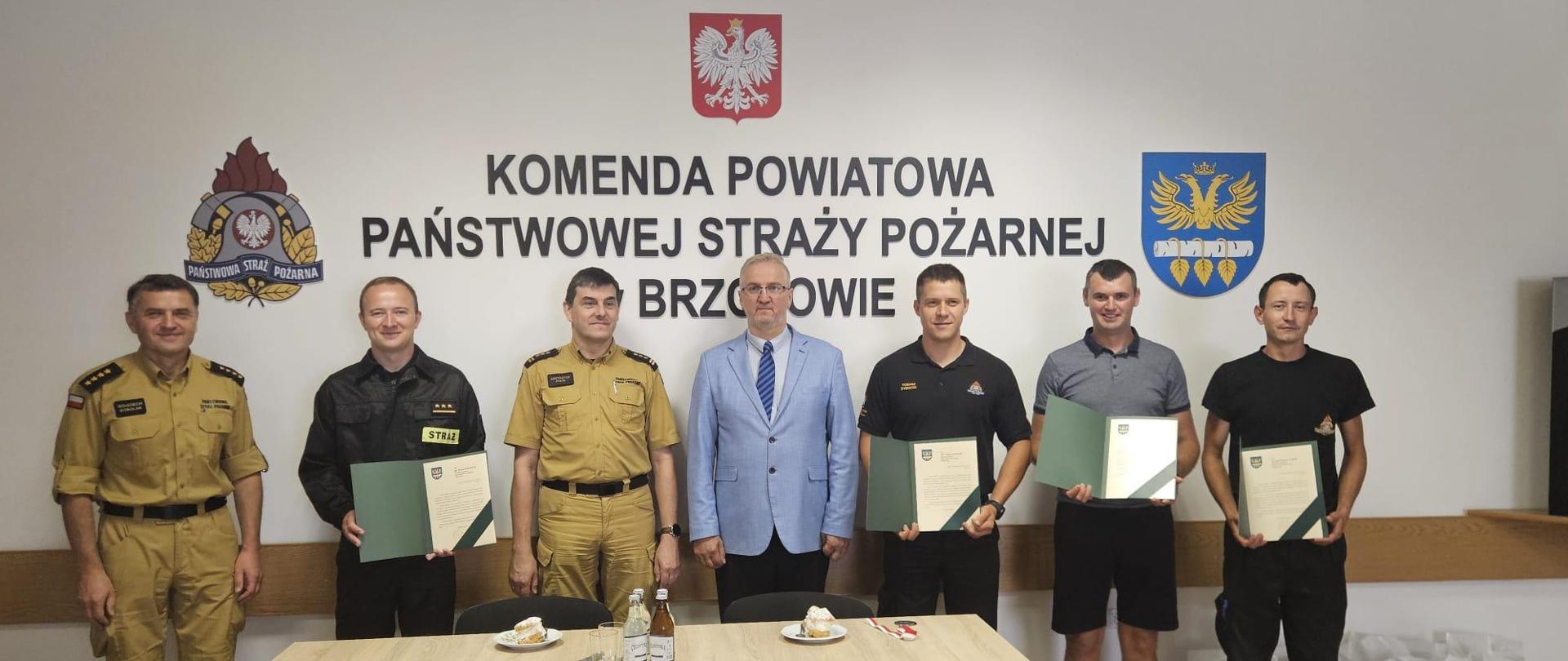 Kolorowa fotografia wykonana w pomieszczeniu. Przedstawia grupę strażaków w strojach służbowych i cywilnych po wręczeniu listów gratulacyjnych przez starostę brzozowskiego i komendanta PSP w Brzozowie.