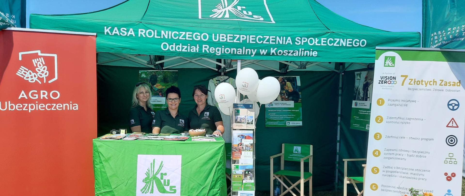 Zielony namiot Oddziału regionalnego KRUS w Koszalinie. Przy stoisku za pulpitem trzy kobiety pracownice OR w Koszalinie, obok ulotki i białe balony, po bokach baner Agro - Ubezpieczenia po lewej i Vision Zero KRUS - po prawej.