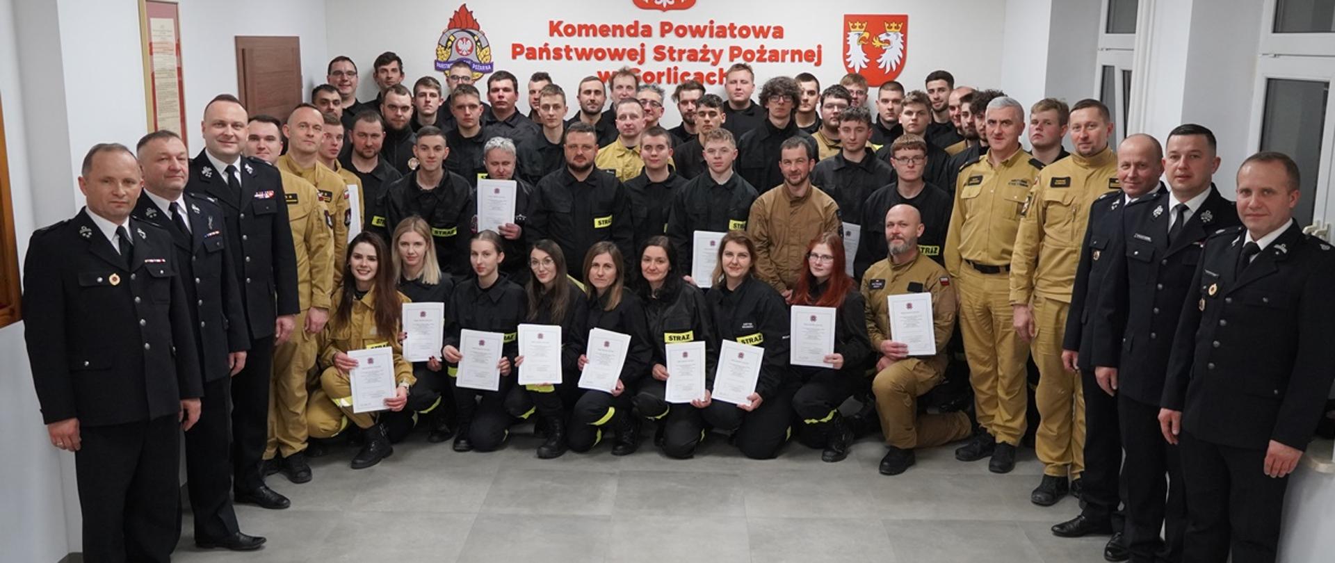 Na zdjęciu w sali konferencyjnej Komendy Powiatowej Państwowej Straży Pożarnej widoczni strażacy Państwowej Straży Pożarnej i druhowie kończący szkolenie podstawowe strażaków ratowników Ochotniczych Straży Pożarnych ubrani w ubrania koszarowe w czarnym kolorze podczas uroczystego zakończenia szkolenia.