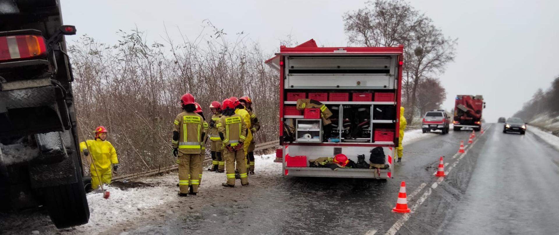 Zdjęcie przedstawia samochody strażackie stojące na drodze, na której miał miejsce wcześniej wypadek. Na zdjęciu widać częściowo tył naczepy samochodu ciężarowego. Strażacy stoją przy samochodzie strażackim. Jeden pas drogi wygrodzony pachołkami. 