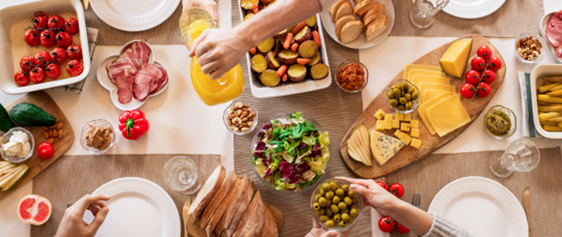 Na zdjęciu znajduje się stół wypełniony żywnością w postaci pieczywa, warzyw, owoców, wartościowych tłuszczy oraz nabiału. Widoczne są również dłonie 3 osób siedzących przy zastawie znajdującej się na stole.