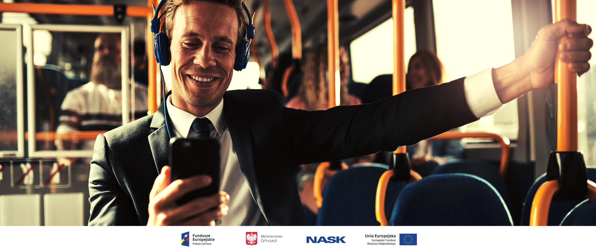 Uśmiechnięty mężczyzna jedzie autobusem. Ma założone słuchawki i wpatruje się w smartfona.
