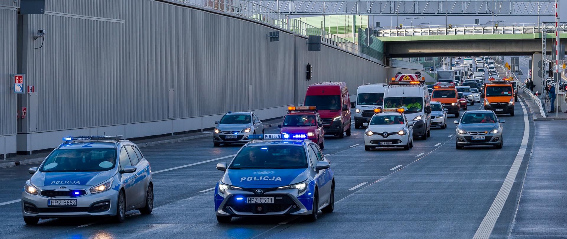 Zdjęcie przedstawia scenę na wielopasmowej drodze z wieloma pojazdami, w tym dwoma radiowozami z migającymi niebieskimi światłami, czerwonym vanem oraz kilkoma innymi samochodami i pojazdami serwisowymi GDDKiA. Wszystkie pojazdy poruszają się za dwoma radiowozami na pierwszym planie.