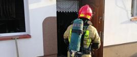 Strażak w ubraniu specjalnym i aparacie ochrony dróg oddechowych zbliża się do drzwi wejściowych kotłowni w której powstał pożar