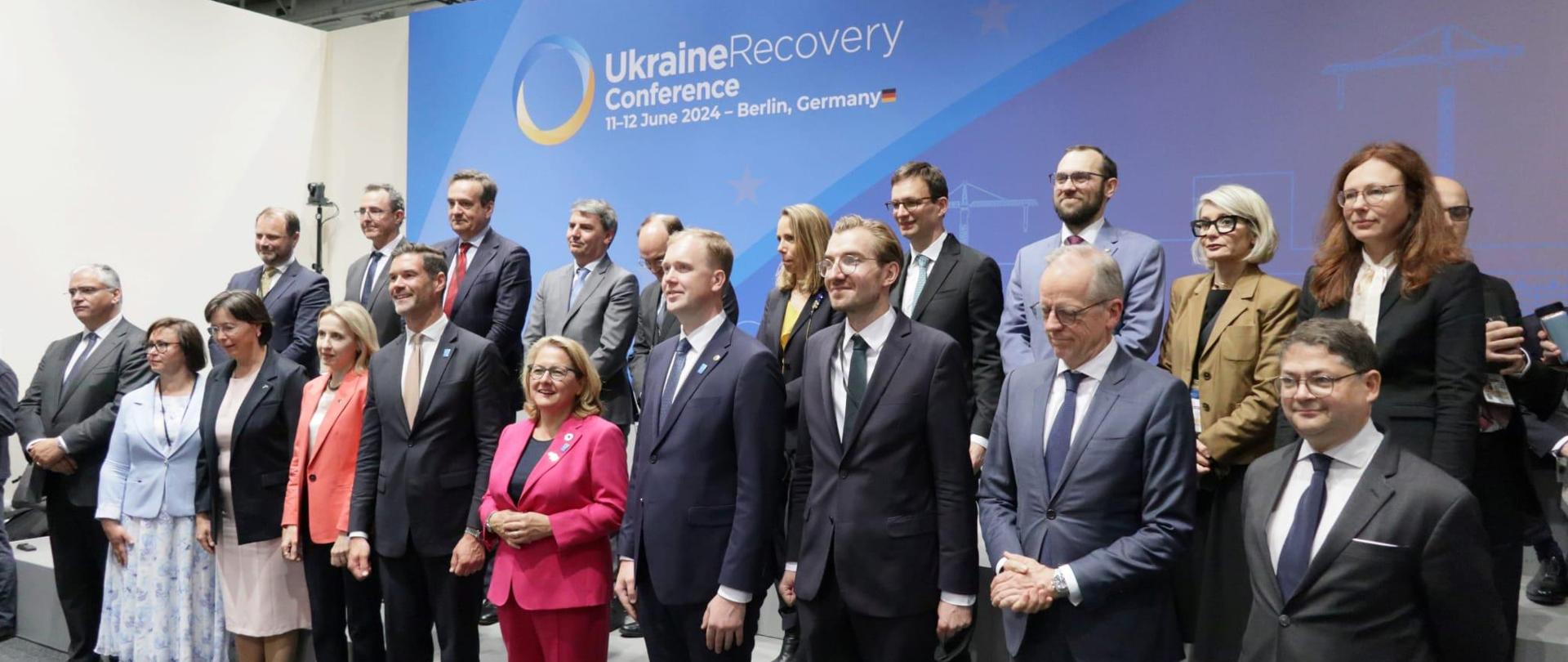 Grupa osób w dwóch rzędach pozuje do zdjęcia. Za nimi ścianka z napisem Ukraine Recovery Conference.