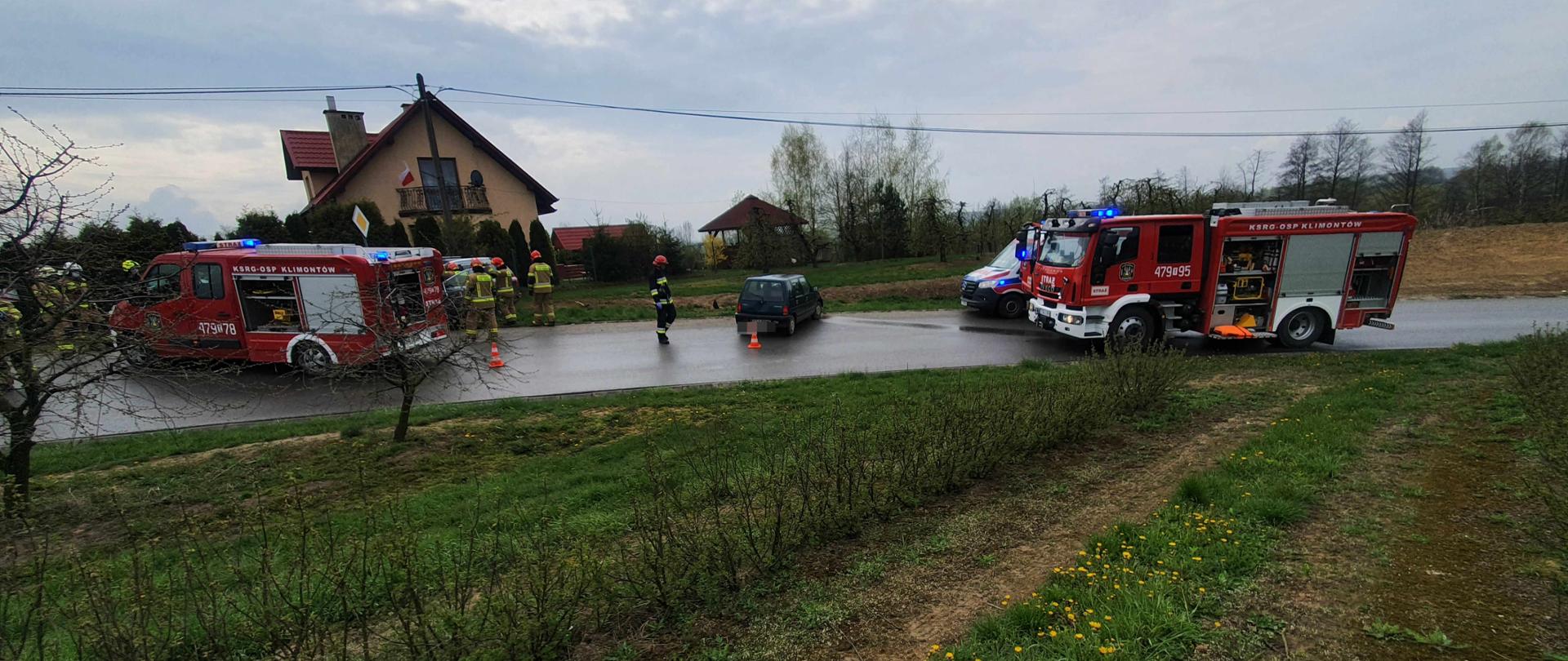 Zdjęcie wykonane w godzinach porannych. Na pierwszym planie widać ustawione samochody służb ratowniczych , oraz strażaków pracujących przy uszkodzonym samochodzie osobowym . Auta ustawione są na drodze asfaltowej zabezpieczone stożkami ostrzegawczymi. W tle widoczny pobliski dom , drzewa , ;linie energetyczne .