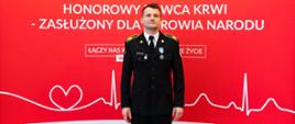 Na zdjęciu widzimy strażaka w galowym stroju na tle biało czerwonej tablicy z napisem "Honorowy dawca krwi - zasłużony dla zdrowia narodu