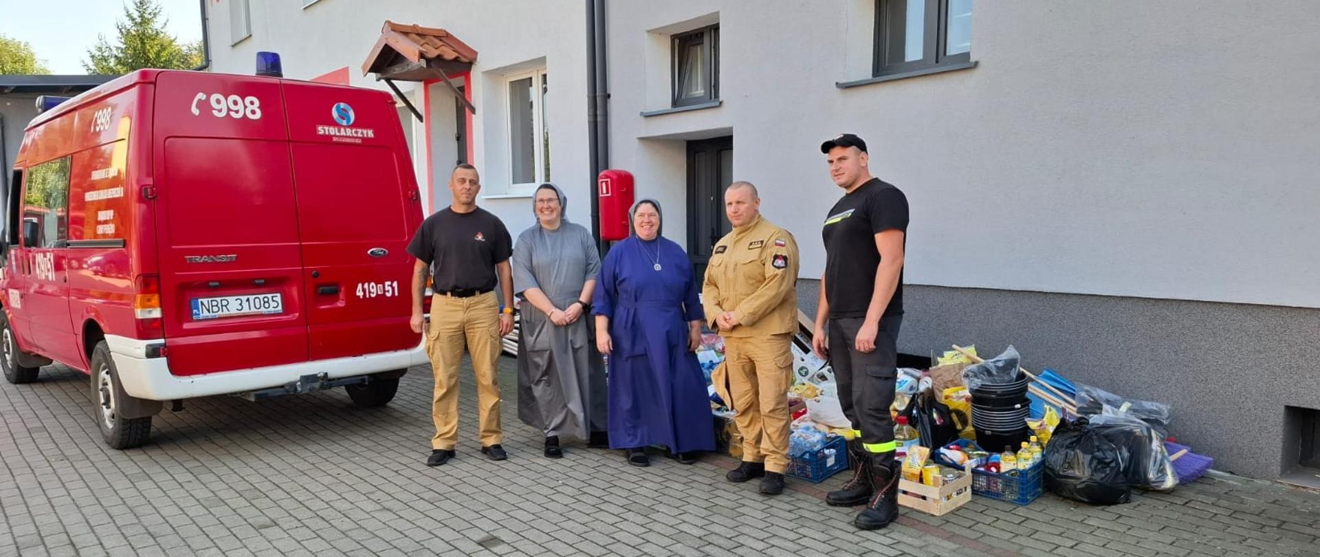 Dwie siostry katarzynki ze strażakami, stoją przy przekazanych darach dla powodzian.