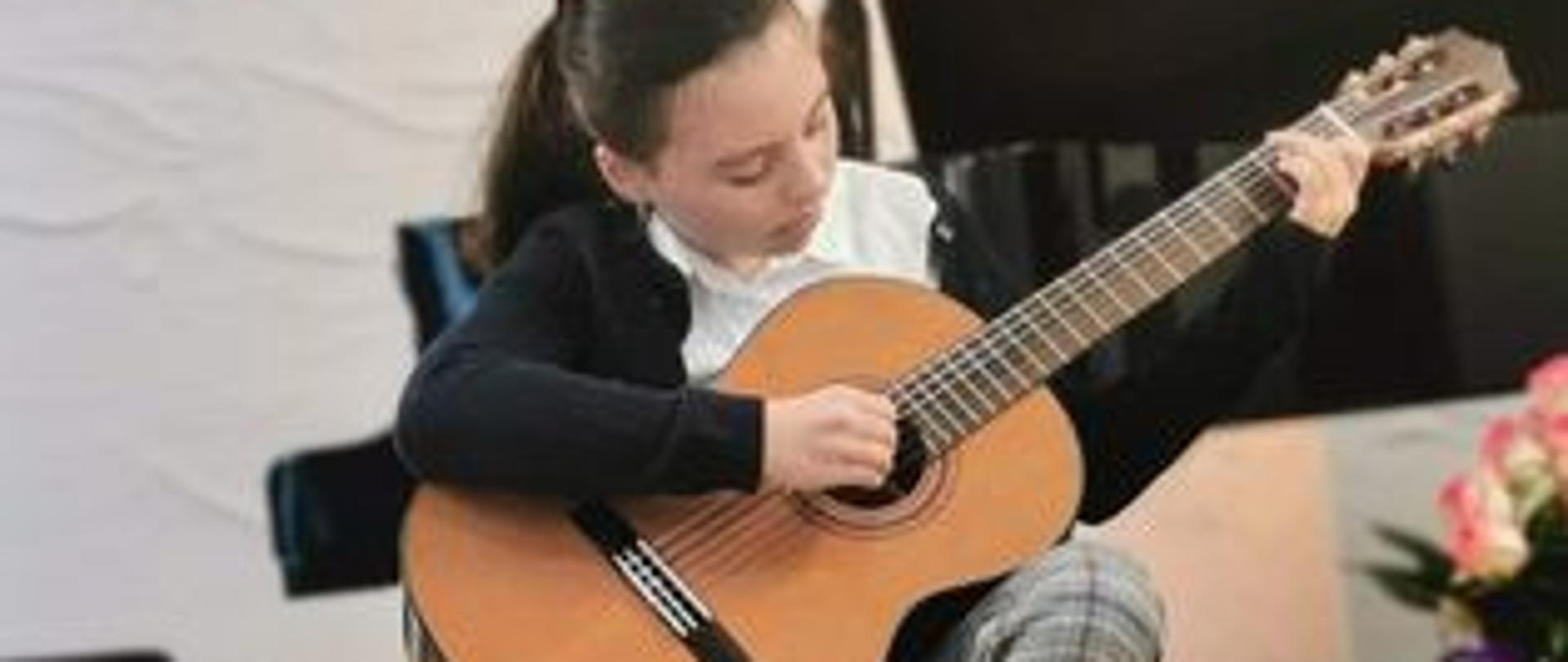 Na zdjęciu znajduje się dziewczynka grająca na gitarze, ubrana w czarny sweter, białą koszulę, spodnie w kratkę ( szaro-niebiesko-białą) i białe buty, gitara ma kolor jasnego drewna , podłoga ma kolor jasnego drewna , w tle czarny fortepian i czarny stołek do fortepianu oraz biała ściana.