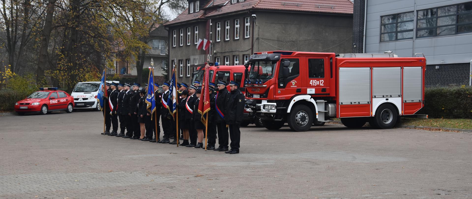 Poczty sztandarowe i strażacy stojący na placu w trakcie uroczystości, w tle widoczne samochody strażackie.