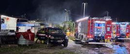 Zdjęcie przedstawia pożar w zakładzie do produkcji papieru. Zdjęcie przedstawia samochody strażackie oraz strażaków. Nad jednym z budynków unosi się dym. 