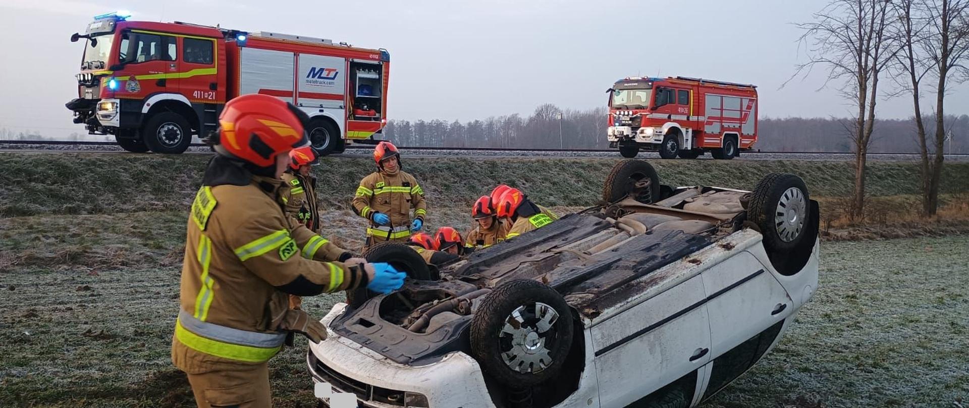 W polu, poza drogą białe auto osobowe na dachu. Przy aucie strażacy w czerwonych hełmach. Dalej na drodze dwa duże samochody strażackie.