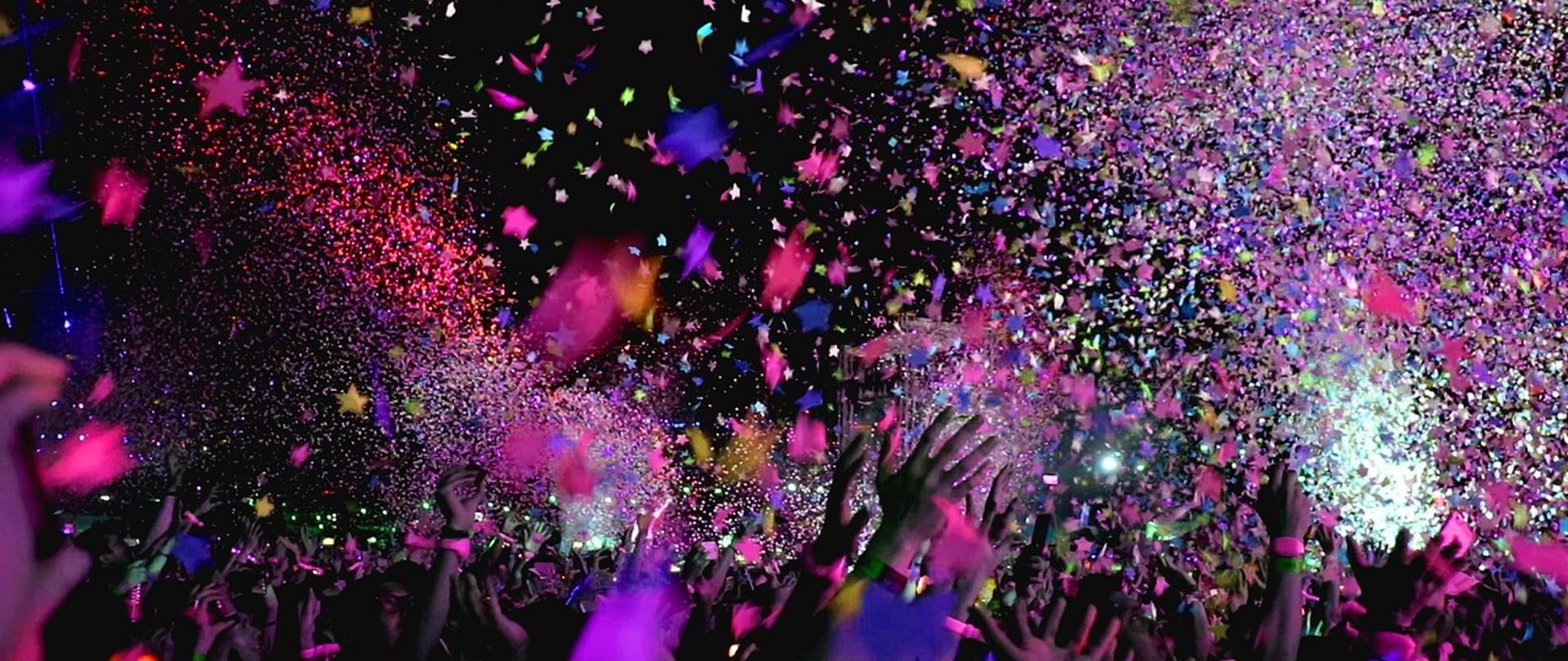 Kolorowe konfetti w tle koncertu, na dole zdjęcie w kolorowym oświetleniu widoczne wyciągnięte do góry ręce. 
