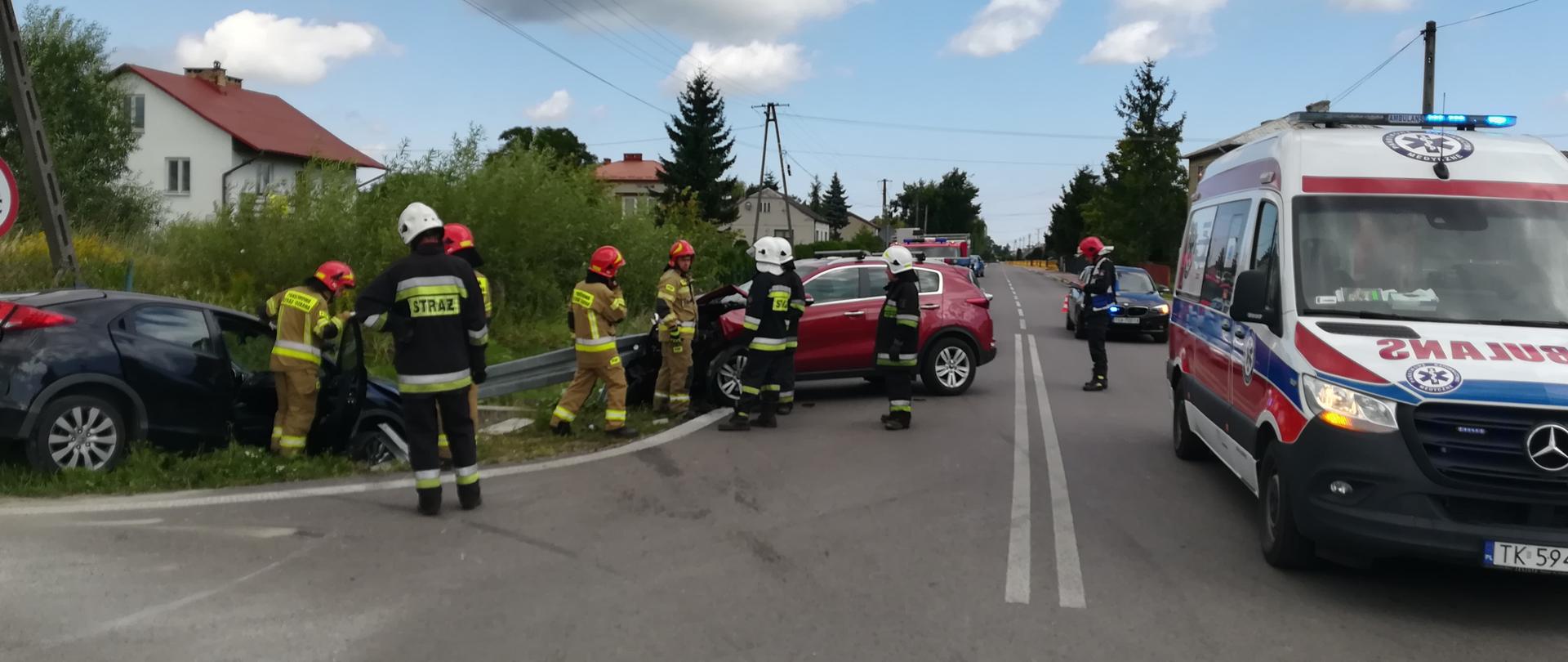 Na pierwszym planie zdjęcia widać dwa rozbite samochody osobowe biorące udział w zdarzeniu i strażaków podczas wykonywania działań . Widać również ambulans Państwowego Pogotowia Ratunkowego i w oddali samochód Policji.